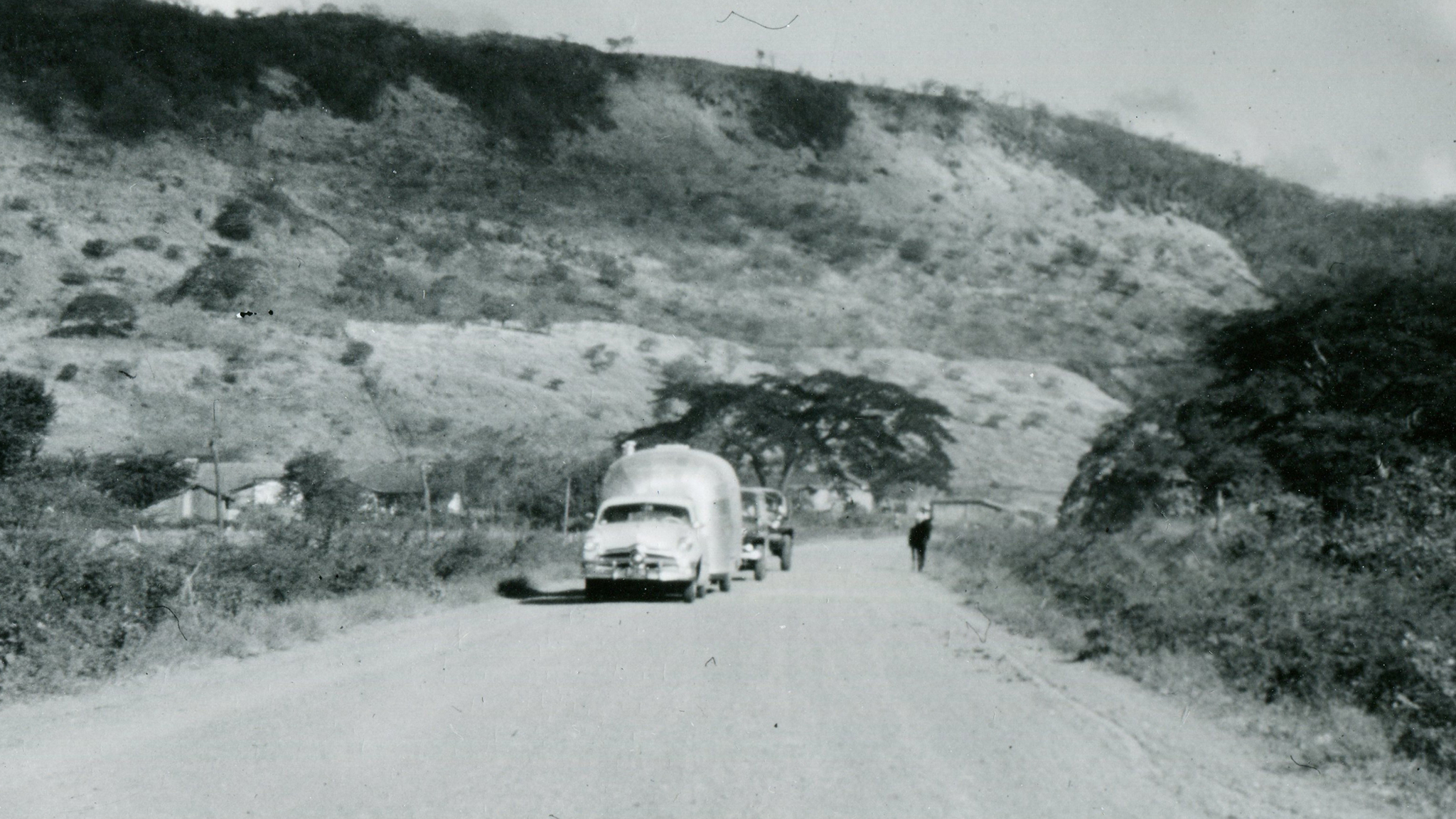 Black-and-white photo of three vintage vehicles driving on a dirt road through a rural, hilly landscape with scattered trees and bushes. A person walks along the road towards the vehicles.