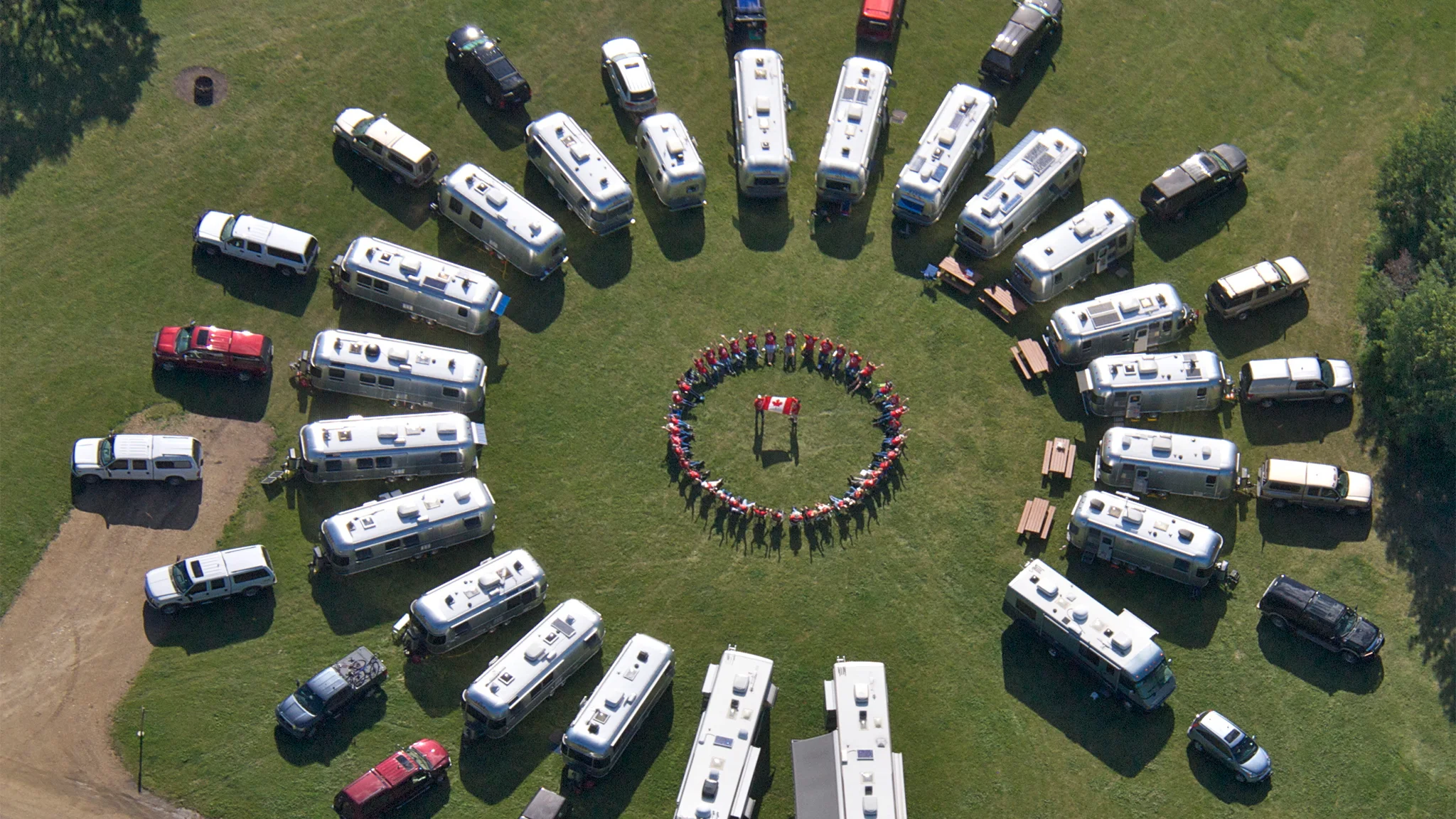 Aerial view of RVs arranged in a circle on grass, surrounding a group of people gathered around a flag in the center.