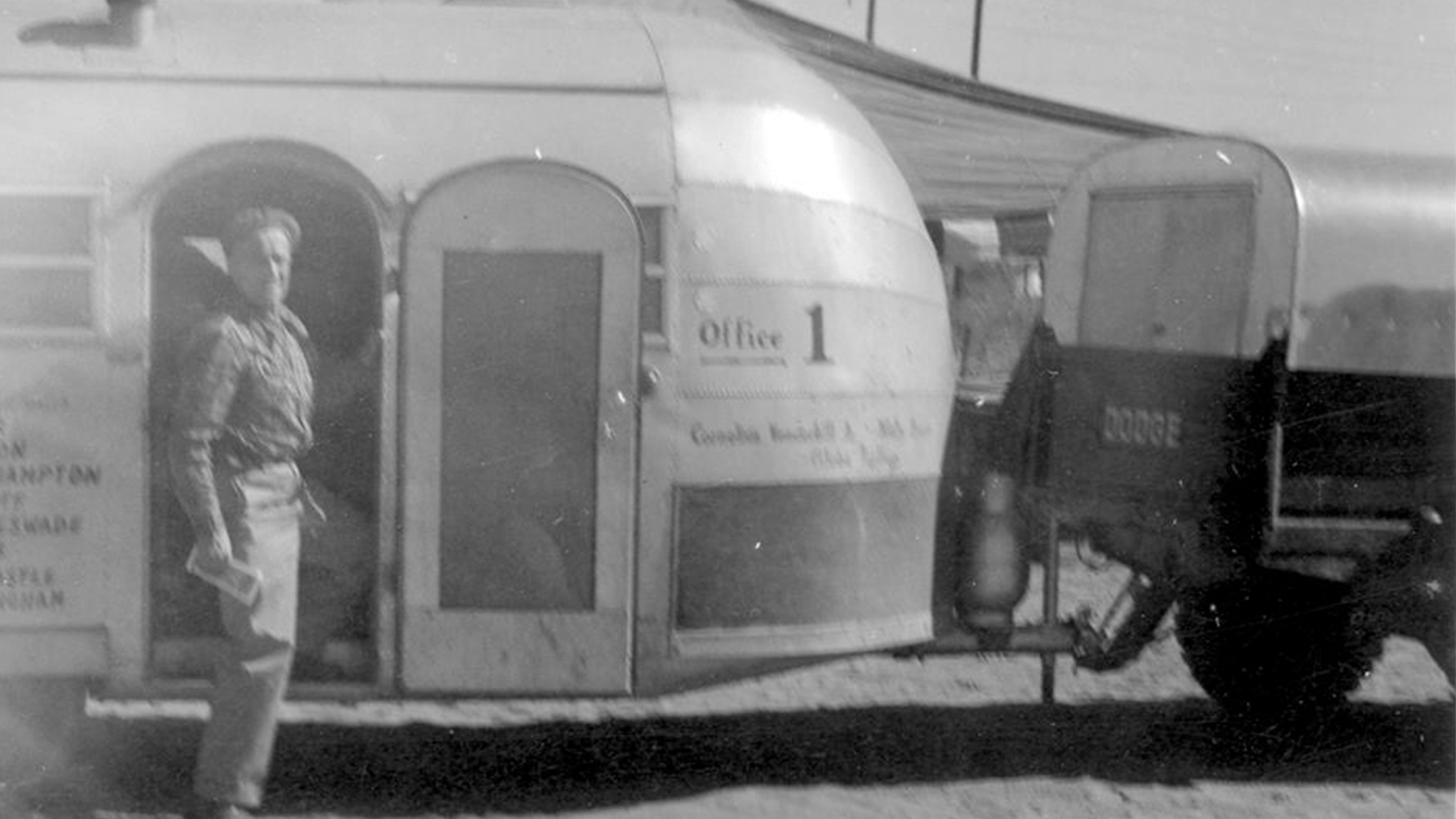 A black and white photo of a person standing beside a vintage travel trailer labeled Office 1, which is hitched to a vehicle. The scene appears to be outdoors in a sunny, possibly rural area.