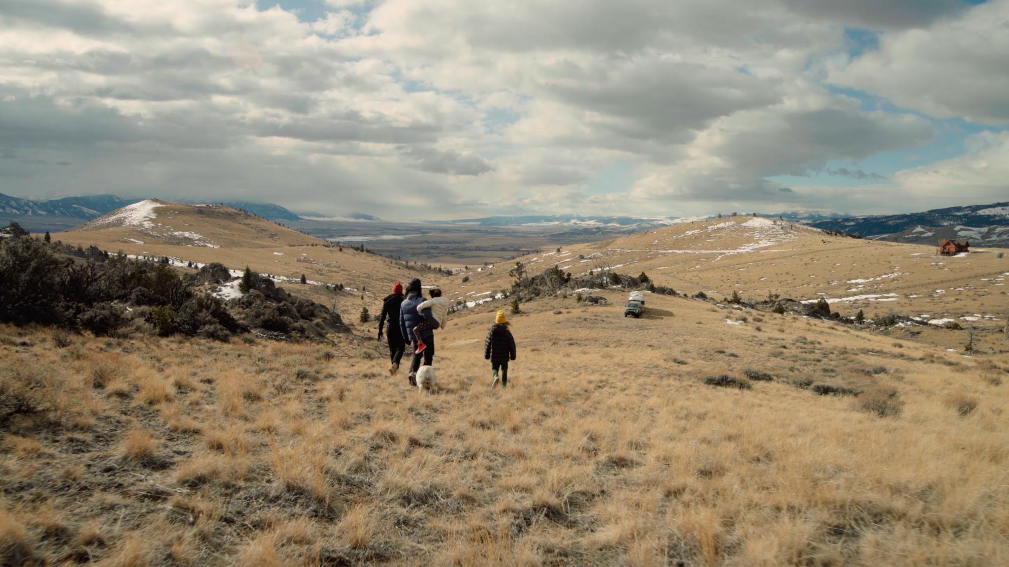 Family with dog walking in a vast, hilly landscape. Snow-dusted hills, cloudy sky, and distant mountains create a serene, expansive view.