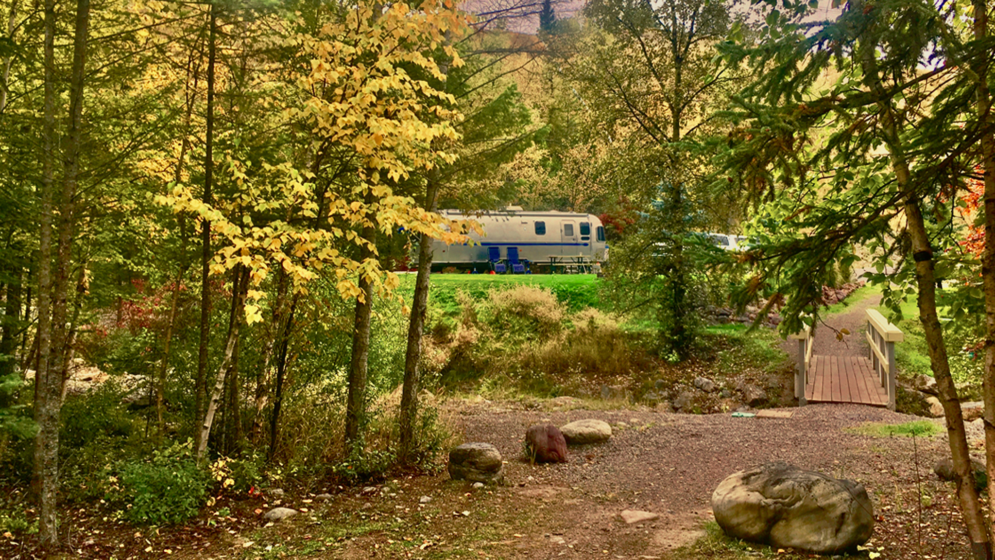 A silver trailer is parked among trees with autumn foliage. A small wooden bridge crosses a rocky path in the foreground, and a blue picnic table is visible near the trailer.