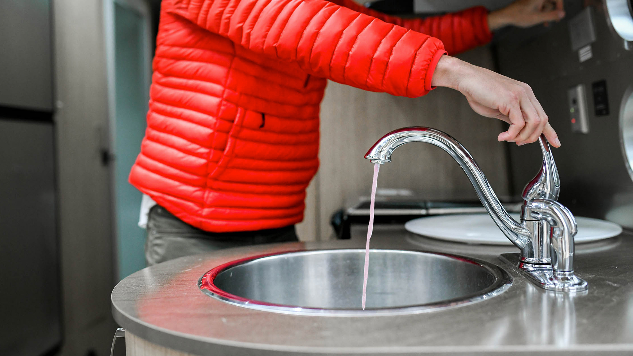 A person in a bright red jacket stands at a modern sink, turning on the faucet with one hand. Water is trickling from the faucet into the round metal basin.