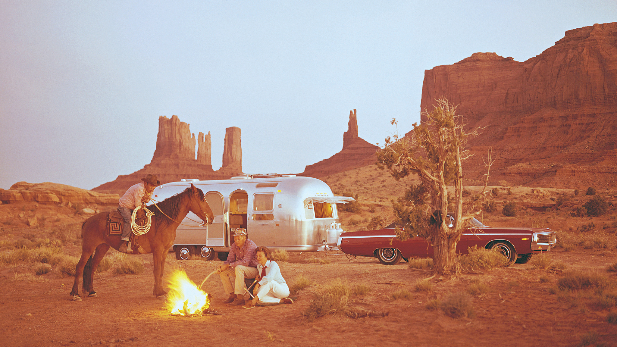 Three people and a horse gather at a campfire in a desert with red rock formations. An Airstream trailer and a classic car are parked nearby, and the scene is bathed in warm, golden light.