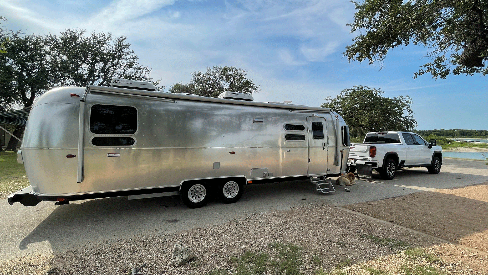 A white pickup truck is hitched to a shiny silver Airstream travel trailer parked near trees and a body of water, with a dog resting on the ground beside the trailer.