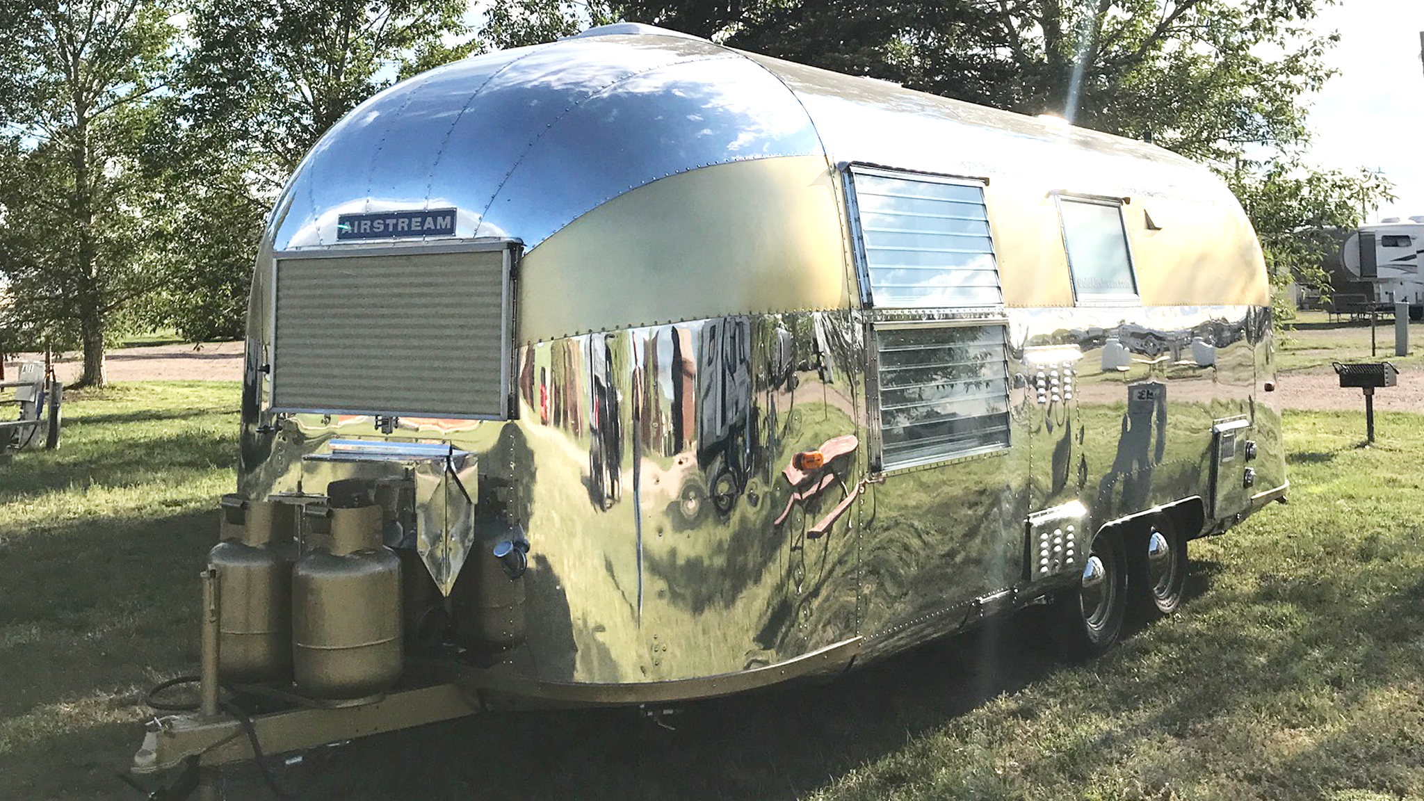 A shiny silver Airstream trailer is parked on grass, surrounded by trees. The trailer has rounded edges, large windows, and two propane tanks mounted at the front. The sky is partly cloudy.