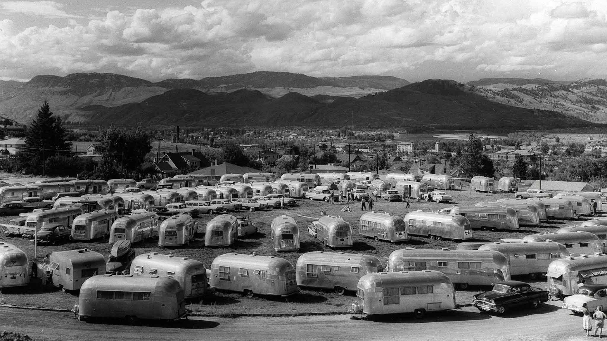 A black and white photo shows a large group of vintage silver trailers parked in a field with mountains in the background and a few cars and people scattered among the trailers under a partly cloudy sky.