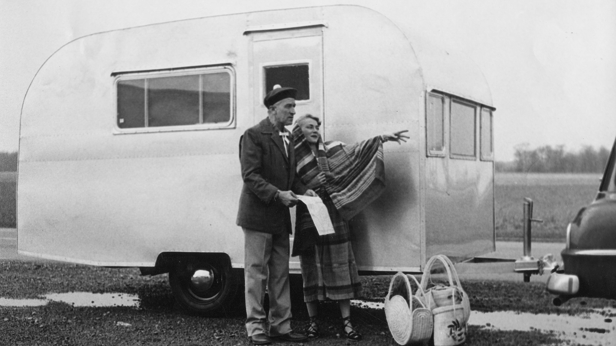 An older man and woman stand beside a vintage travel trailer. The woman points into the distance while the man holds a map. Two baskets are on the ground nearby, and part of a car is visible on the right.