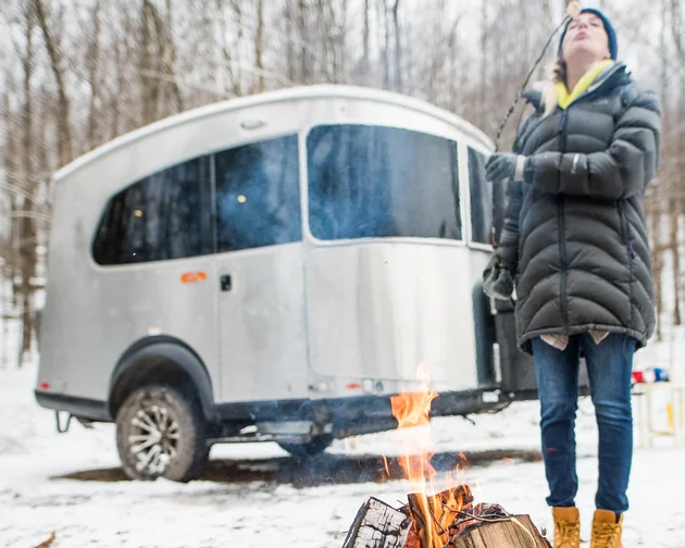 Person in a winter coat toasts a marshmallow over a campfire near a silver camper in a snowy forest.