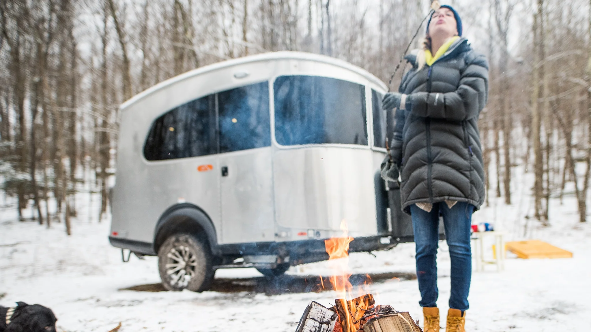 Person in a winter coat toasts a marshmallow over a campfire near a silver camper in a snowy forest.