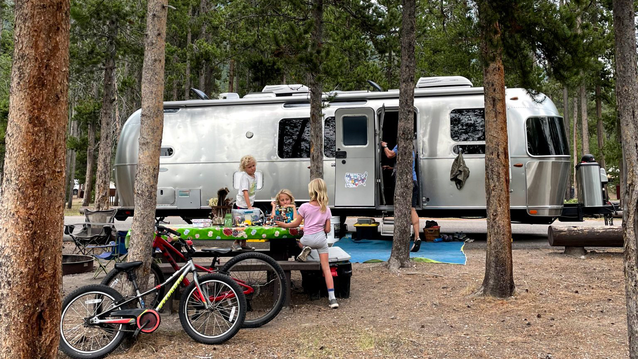 A silver Airstream trailer is parked in a wooded campsite. Two children and an adult sit at a picnic table with a green tablecloth, while another adult stands by the trailer. Bicycles and camping gear are scattered around.