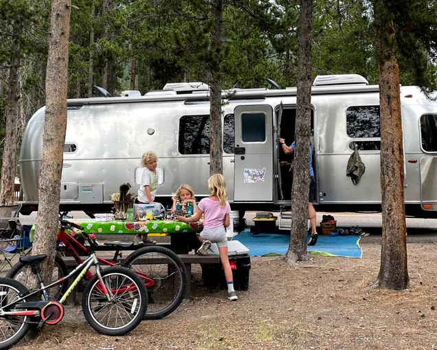 Kids sitting on a picnic table and playing outside of their Airstream travel trailer while they camp.|Fajitas being cooked over a fire.|Two people sitting at a table cooking fish that they just caught.|Vegetables cooking on a grill over a fire.|Pasta noodles and sauce being cooked on a grill.|A plate of scones and a cup of coffee sitting on a table.|Four kids sitting at the table in an Airstream travel trailer eating dinner while they camp.|Three kids sitting at the table in their Airstream travel trailer playing go fish at night with a night light turned on.|A kid cooking fried rice on a grill while he camps in his Airstream Travel Trailer|Two kids on a scooter playing in the dirt while they boondock camp in their Airstream travel trailer|A little kid cooking french toast on a grill next to the Airstream travel trailer while a family camps.|Four kids watching a geyser while they camp.|Kids sitting on a picnic table and playing outside of their Airstream travel trailer while they camp.