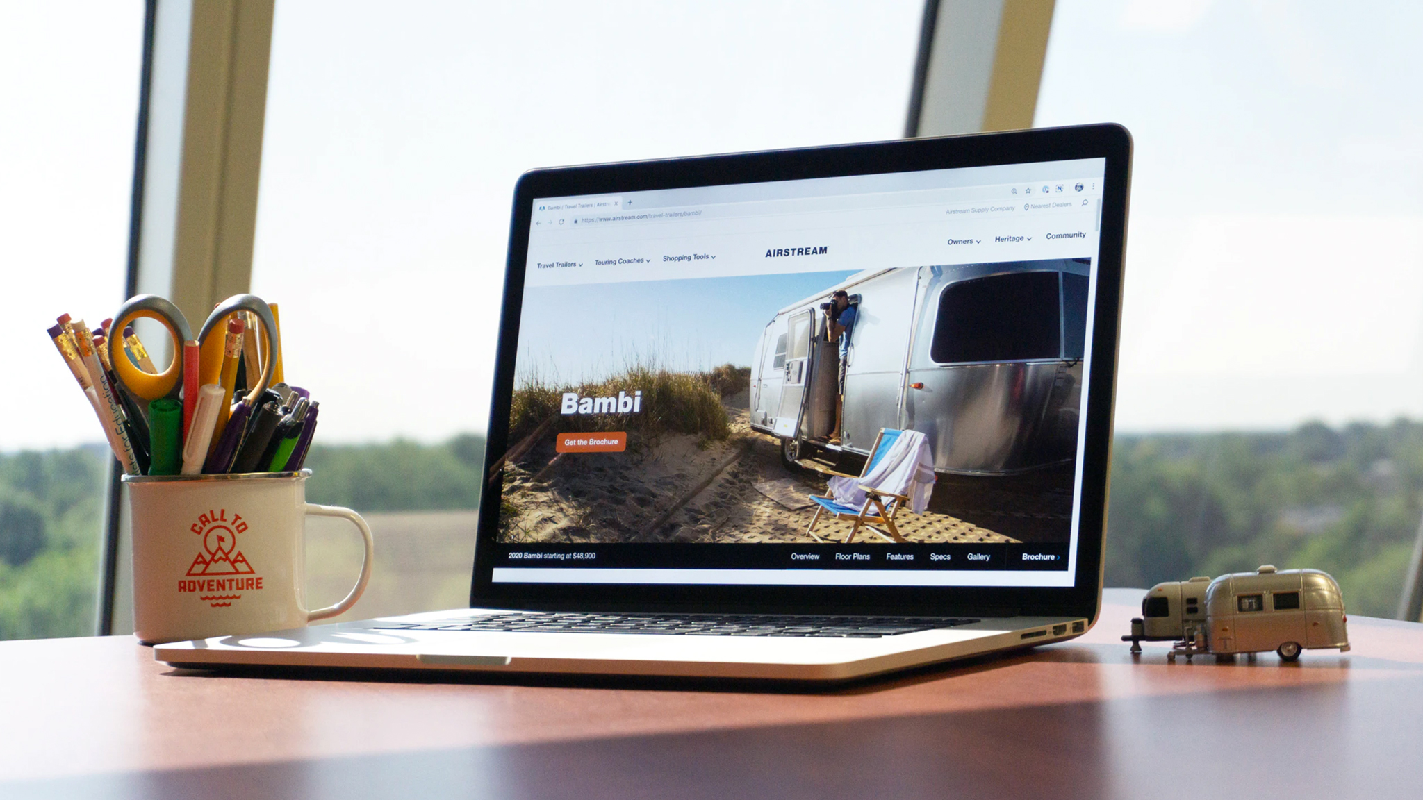 A laptop on a desk displays a website featuring an Airstream trailer. Next to the laptop is a mug filled with pens and scissors, and a small model trailer. Large windows reveal a green, blurry outdoor view.
