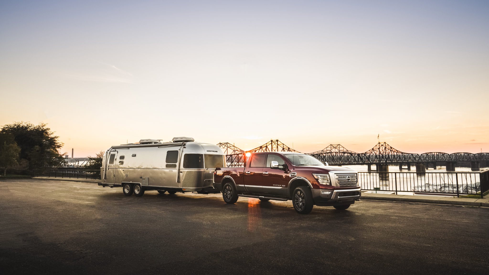 A red pickup truck towing a silver travel trailer is parked by a waterfront at sunset, with a metal bridge and calm water in the background.