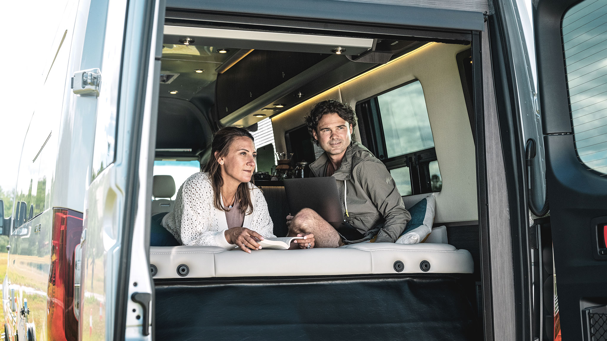 A couple sits in the back of a camper van; the woman holds a book, and the man uses a laptop, both appear relaxed and content.