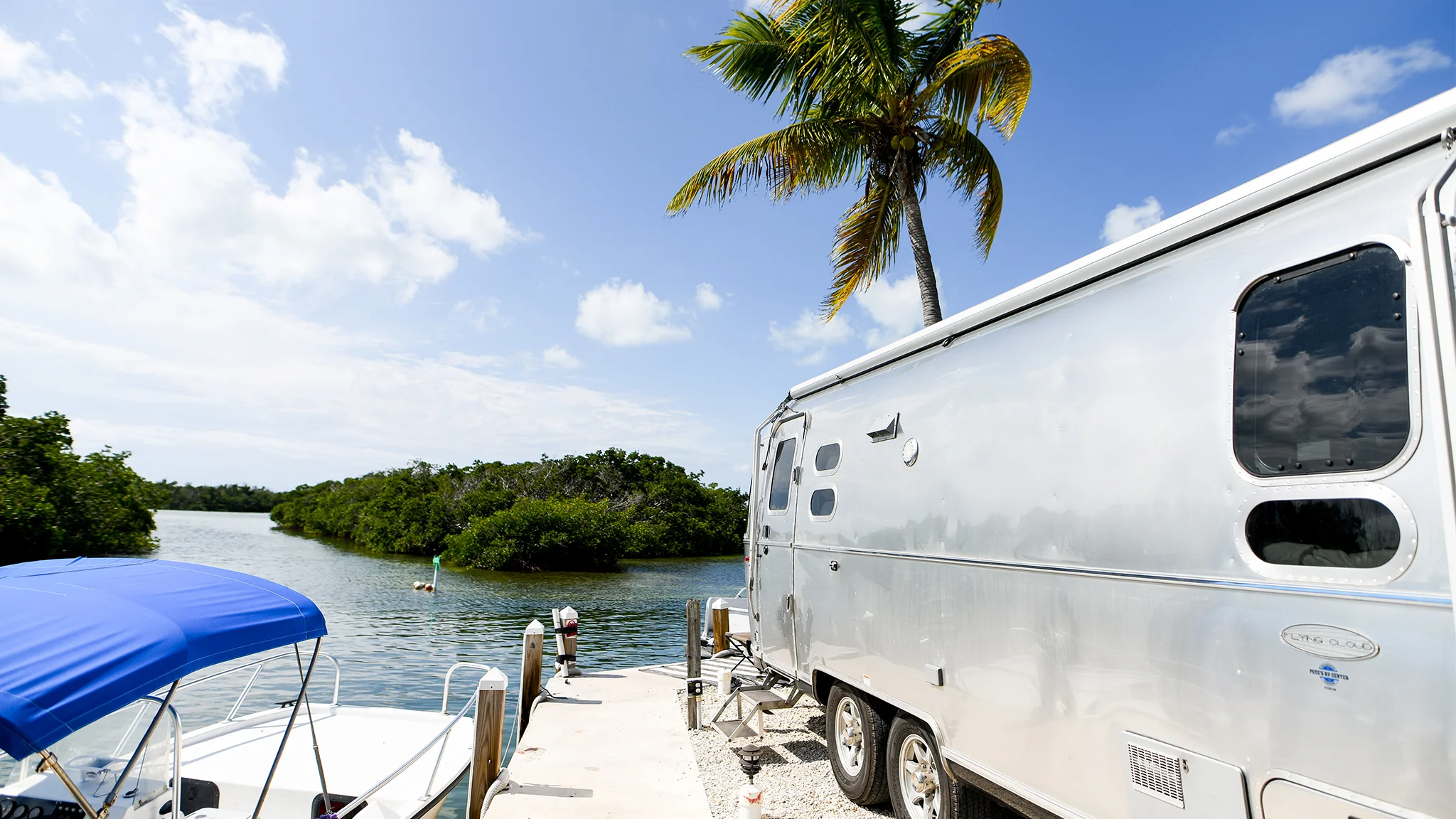 A shiny silver Airstream trailer is parked next to a dock with a blue-and-white boat, palm trees, and water under a bright, partly cloudy sky.