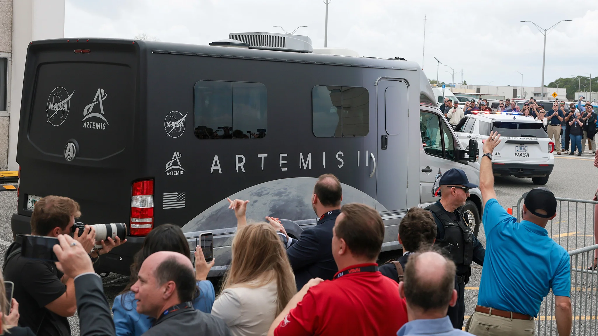 A crowd watches and photographs the NASA Artemis II vehicle leaving a facility, escorted by security, with logos visible on the van.