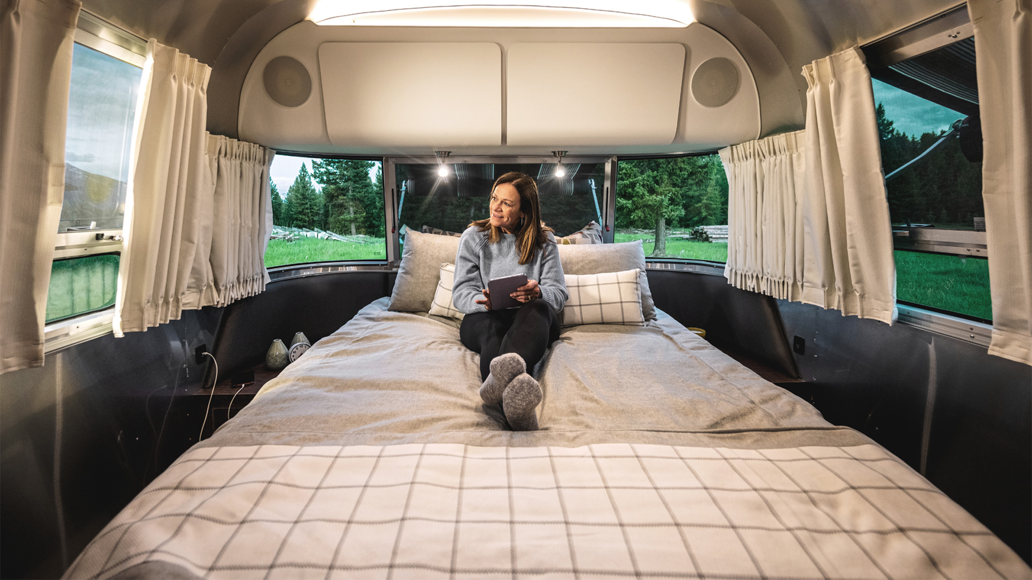 A woman camper sitting on the bed looking at her tablet in her Airstream Globetrotter travel trailer.|Wonderlog logo|recreation-gov-app|togo-rv-app|maps-me|USA-Rest-Stops|iExit-app|gas-buddy-app|campendium-app|KOA-app|All-Stays-app|FreeRoam-app|The-Dyrt-App|Reserve-America|Harvest-Hosts-App|HipCamp-App|RVParky|iOverlander-App|Seek-app|Merlin-Bird|allTrails-app|Hiking-Project-App|Roadtrippers-App|WiFi-Map-App|Open-Signal-App|A woman camper sitting on the bed looking at her tablet in her Airstream Globetrotter travel trailer.