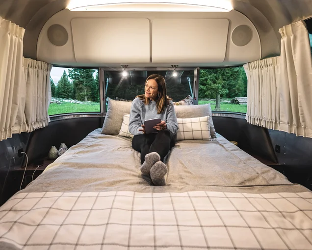 A woman camper sitting on the bed looking at her tablet in her Airstream Globetrotter travel trailer.|Wonderlog logo|recreation-gov-app|togo-rv-app|maps-me|USA-Rest-Stops|iExit-app|gas-buddy-app|campendium-app|KOA-app|All-Stays-app|FreeRoam-app|The-Dyrt-App|Reserve-America|Harvest-Hosts-App|HipCamp-App|RVParky|iOverlander-App|Seek-app|Merlin-Bird|allTrails-app|Hiking-Project-App|Roadtrippers-App|WiFi-Map-App|Open-Signal-App|A woman camper sitting on the bed looking at her tablet in her Airstream Globetrotter travel trailer.