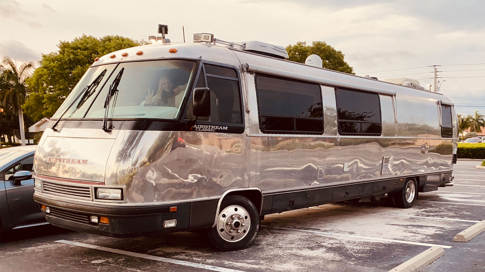 A shiny silver Airstream motorhome is parked in a lot under a partly cloudy sky, surrounded by palm trees and cars, with a person visible in the driver’s seat.