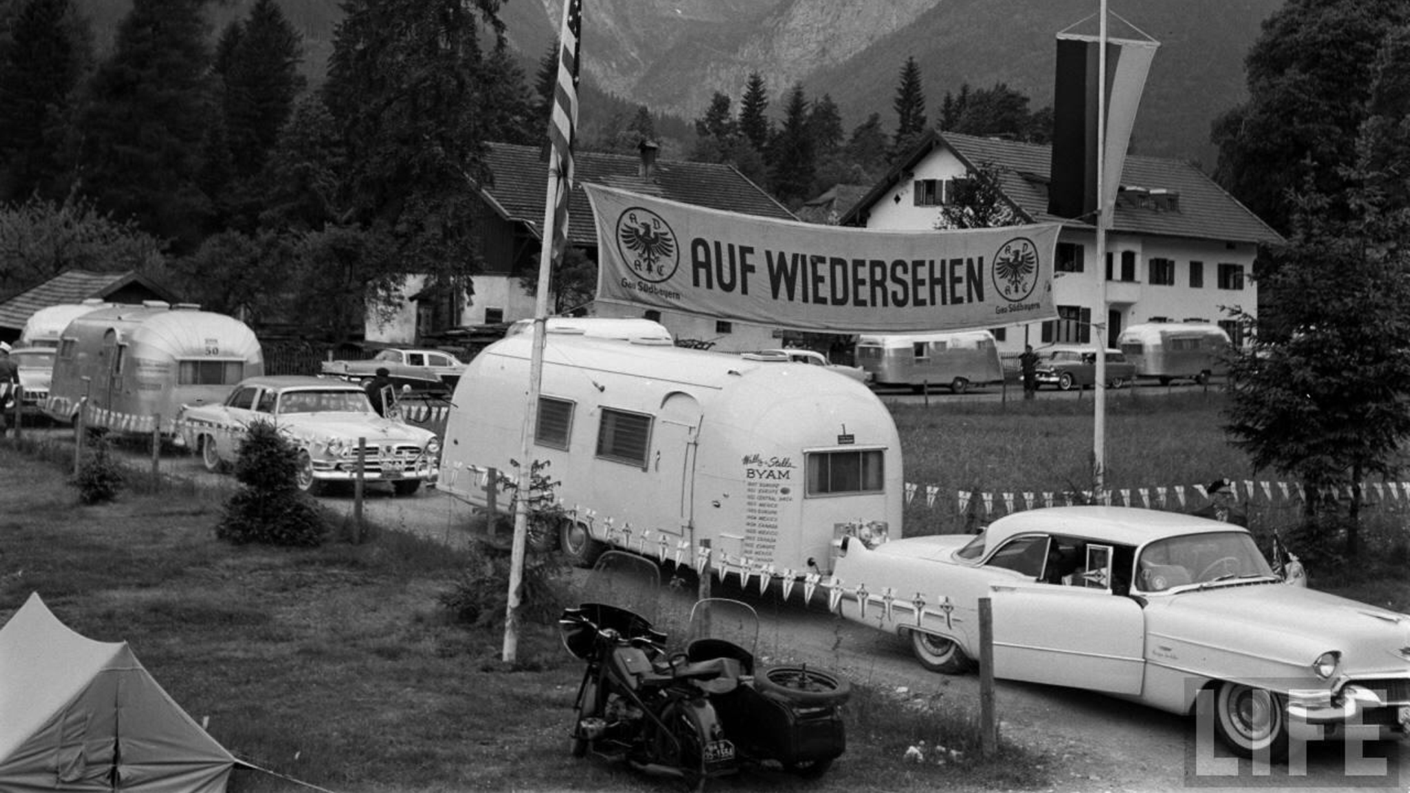 A vintage caravan parked in a scenic area with mountains and trees. A sign reads "AUF WIEDERSEHEN." Classic cars and a motorcycle are nearby.