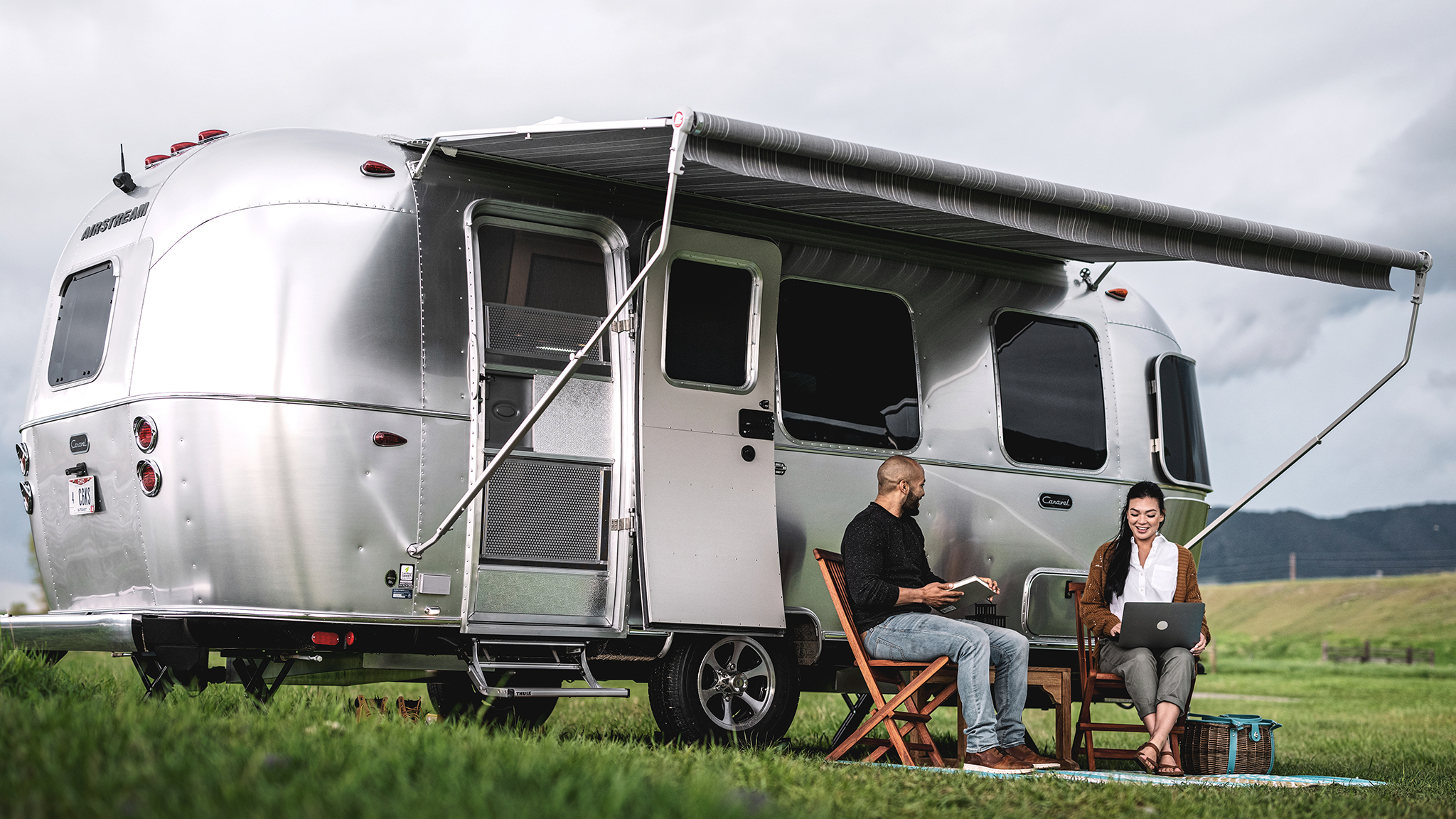 A man and woman working on laptops sit outside a silver camper van with an awning, on a grassy field with hills in the background.