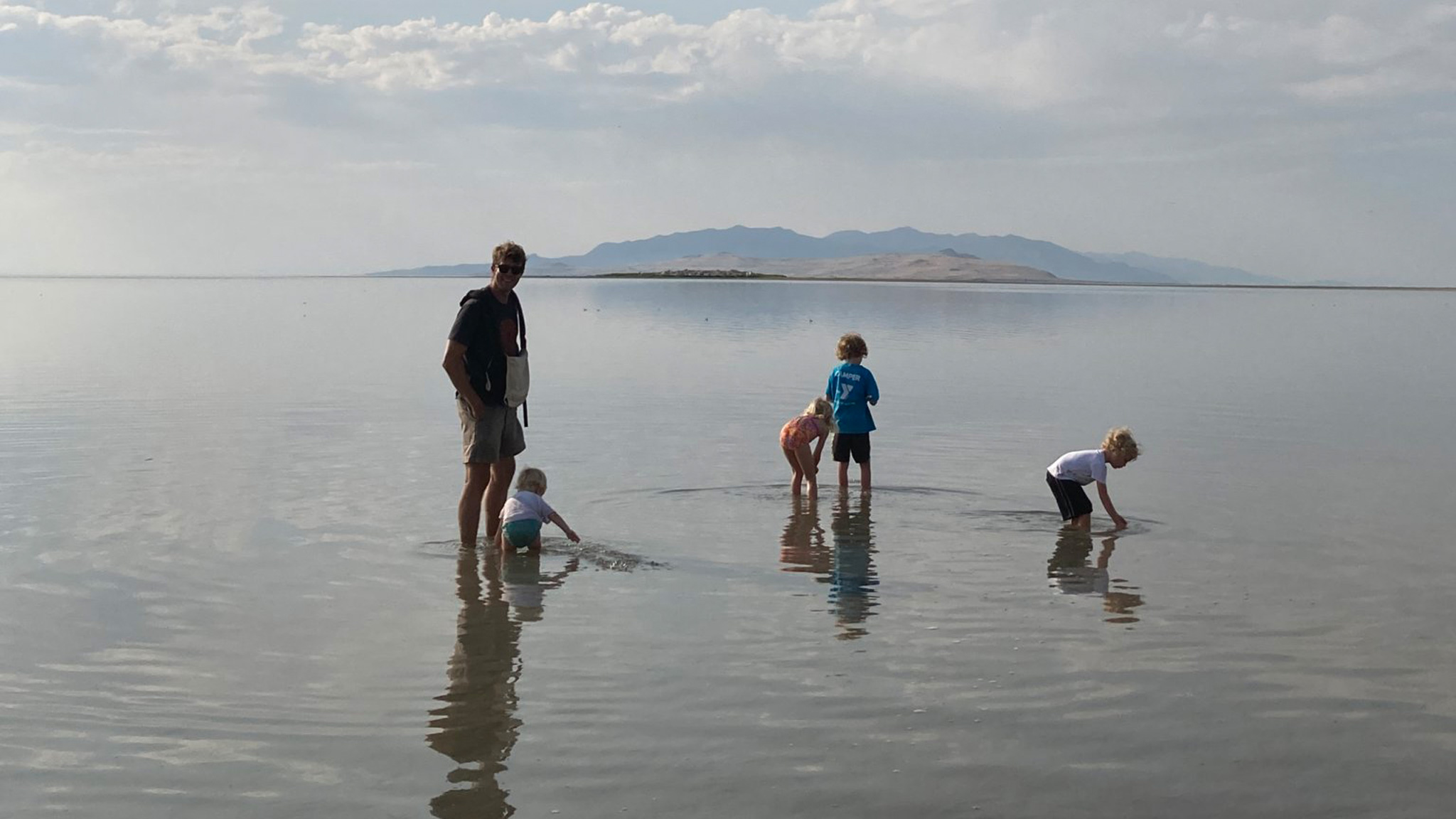 A man and four children wade in shallow water, bending down to explore. An island and mountains are visible in the distance under a cloudy sky.