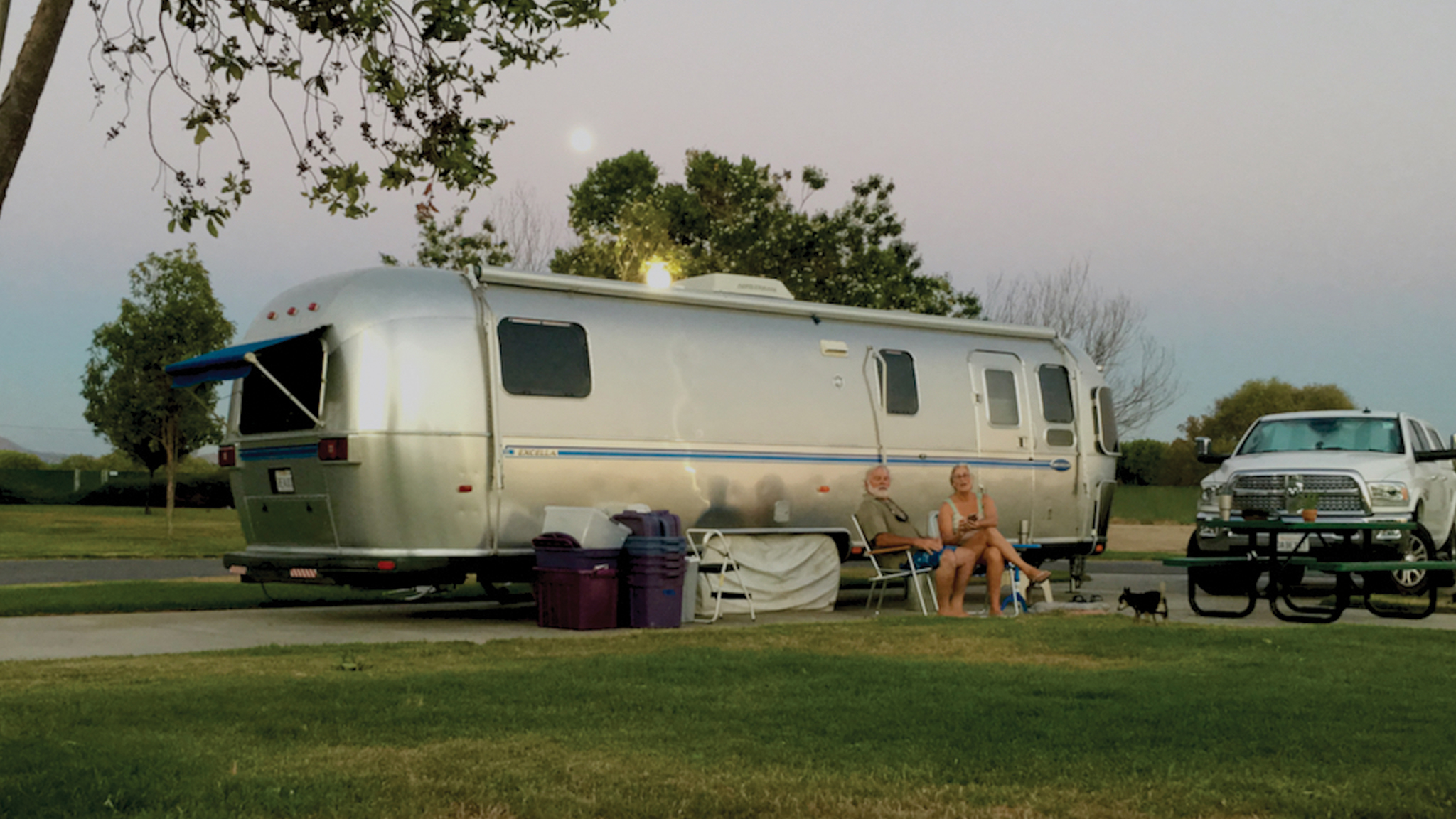 Two people sit outside a silver Airstream trailer at a campsite, with chairs, a table, a small dog, and a white truck parked nearby on grass under a twilight sky.