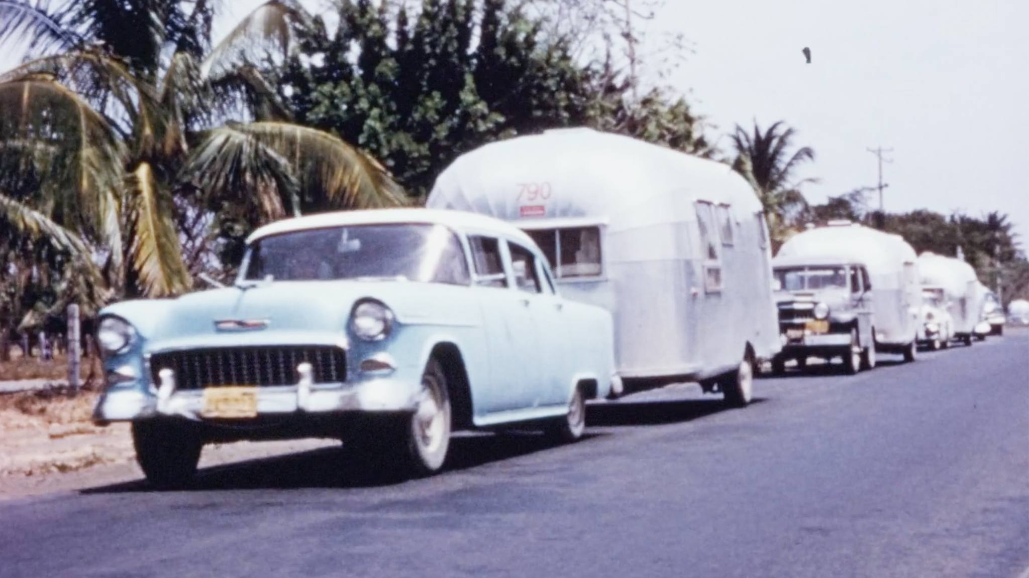 A vintage light blue car towing a shiny silver trailer is parked on a road lined with palm trees, followed by more cars and trailers under a clear sky.