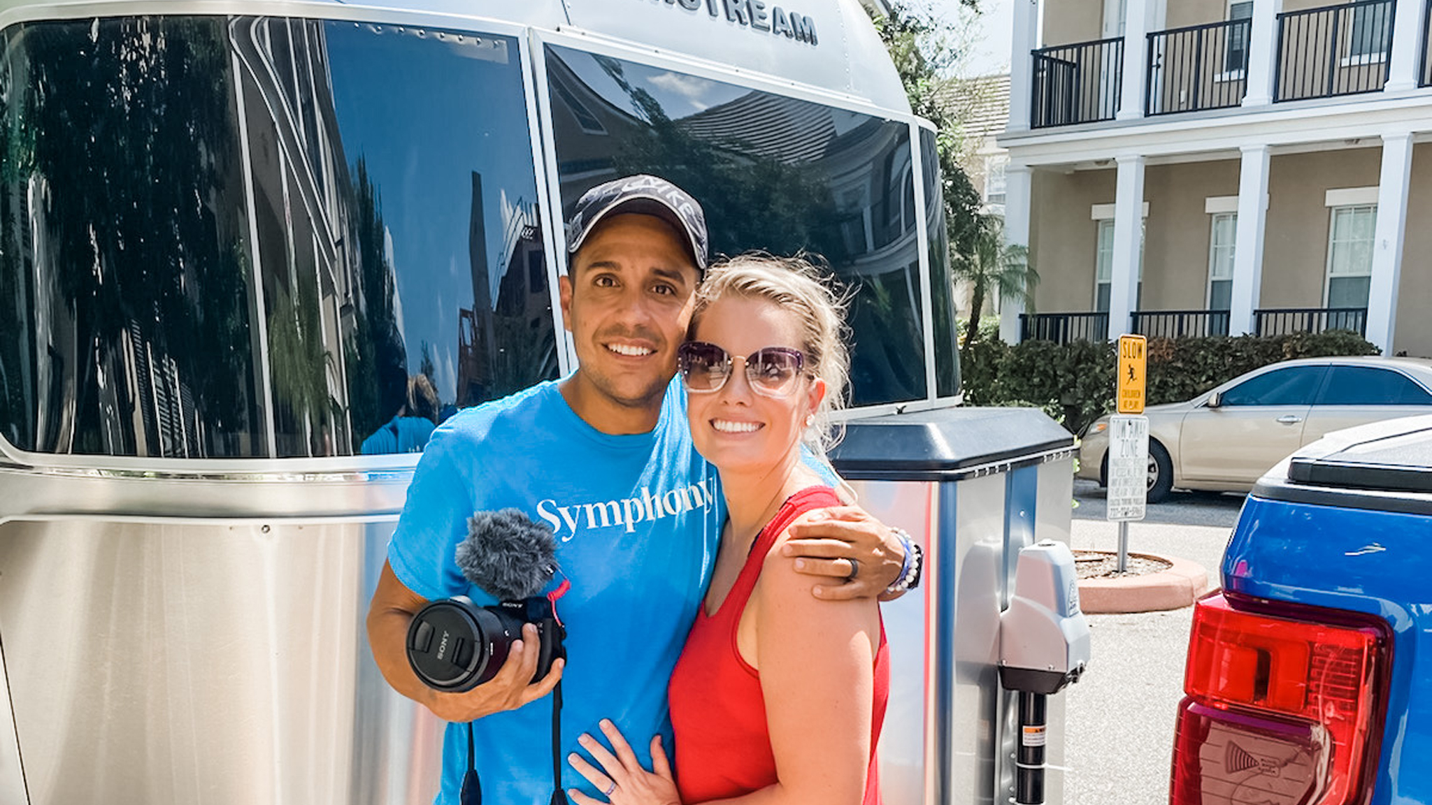 A smiling man and woman pose together outside next to a shiny Airstream trailer. The man holds a camera and microphone, and the woman wears sunglasses and a red tank top. Buildings and a blue truck are in the background.