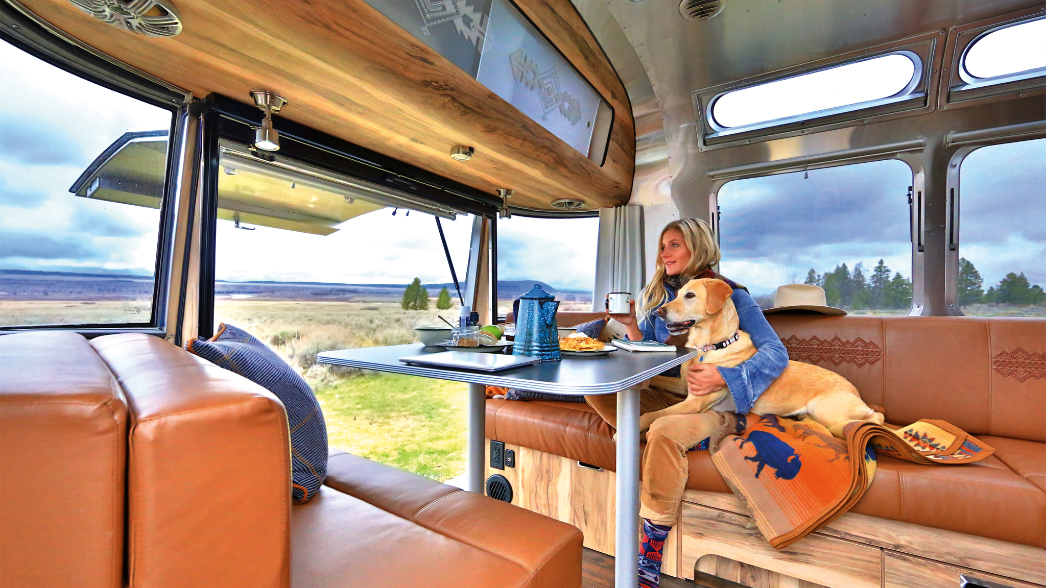 A woman sits inside a camper van at a table with food, accompanied by a dog on her lap. Large windows reveal an open landscape with cloudy skies and distant trees. The interior has brown leather seats and a cozy atmosphere.