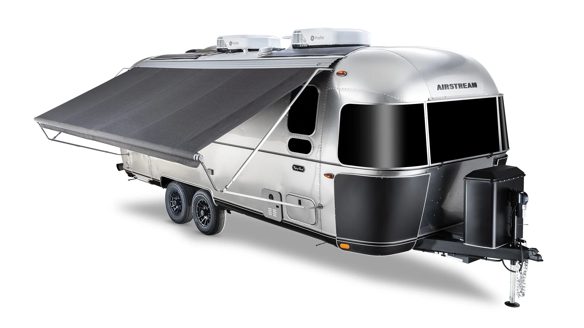 A silver Airstream trailer with a black retractable awning extended, two air conditioning units on top, resting on dual axles.
