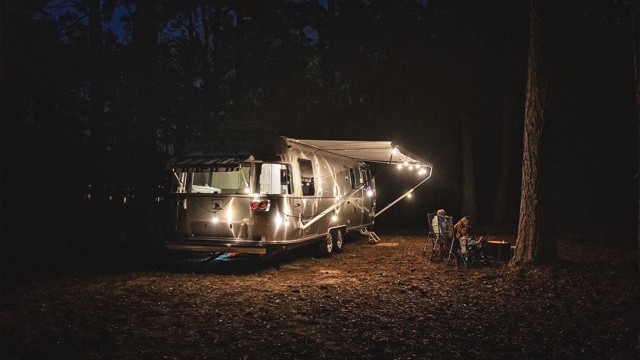 A camper trailer festooned with string lights is parked in a dark, wooded area. Two people sit by a small fire nearby.
