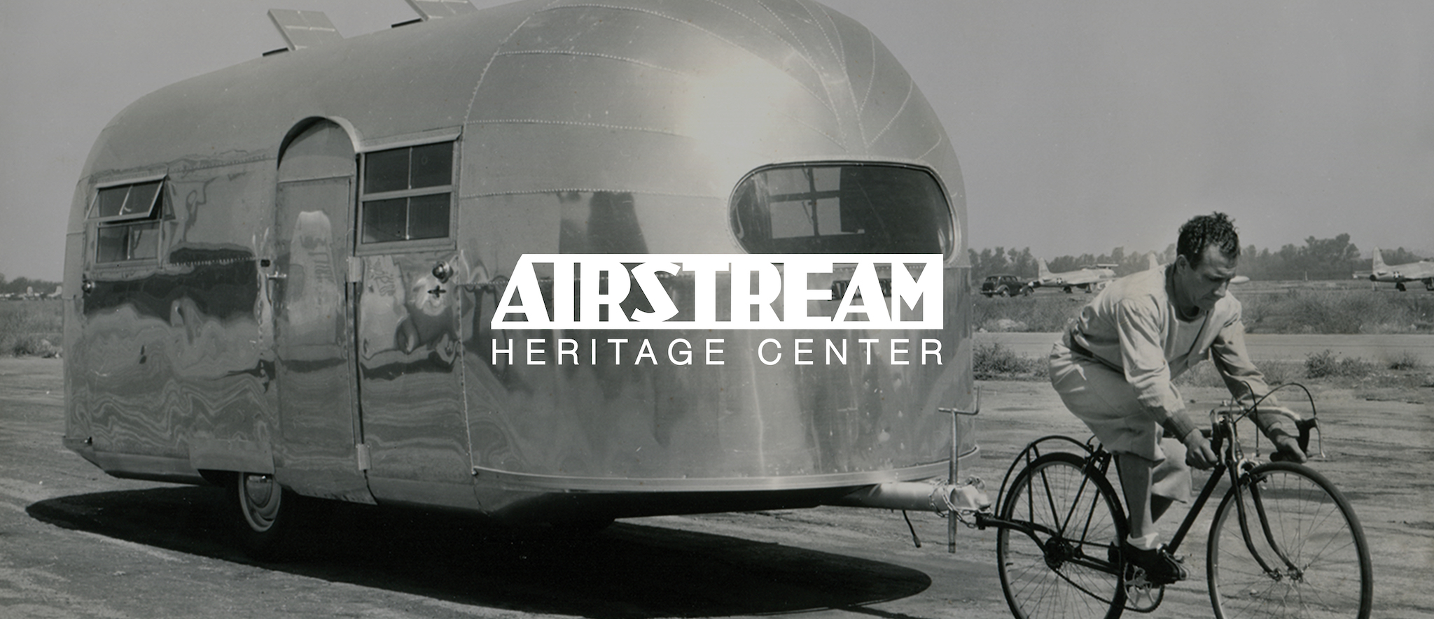 A man in a suit rides a bicycle towing a shiny, vintage Airstream trailer; the image is in black and white with the text “Airstream Heritage Center” overlaid.