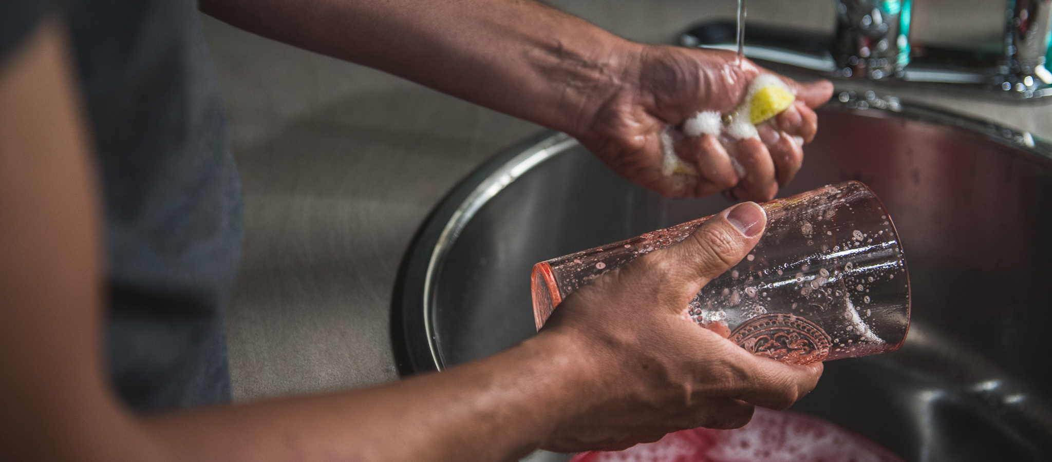 A person is washing a soapy glass cup under running water at a kitchen sink, using a yellow sponge.
