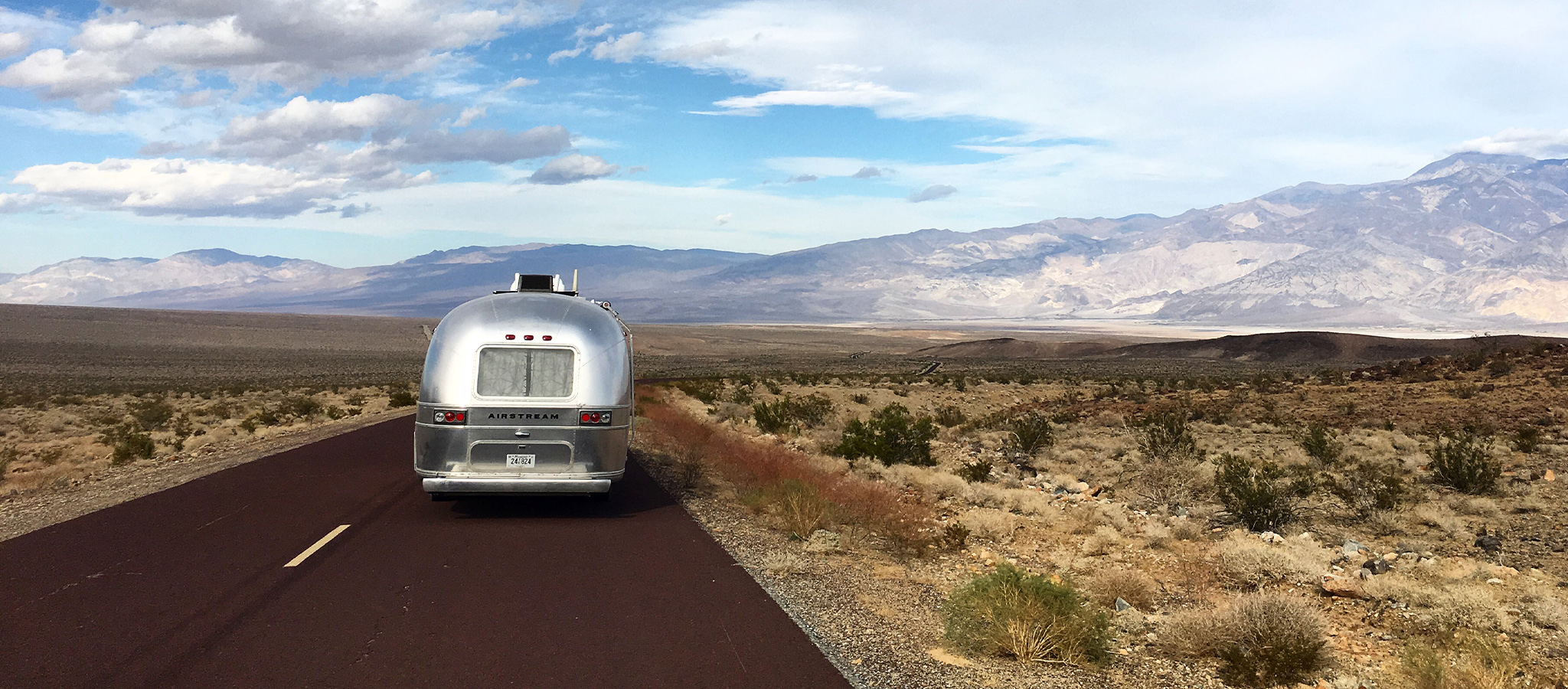 A silver Airstream trailer drives down an empty two-lane road through a vast desert landscape, with mountains and blue sky with scattered clouds in the background.