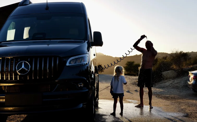 A man and a child stand beside a black van at sunset, with the man using a hose, likely for an outdoor shower.