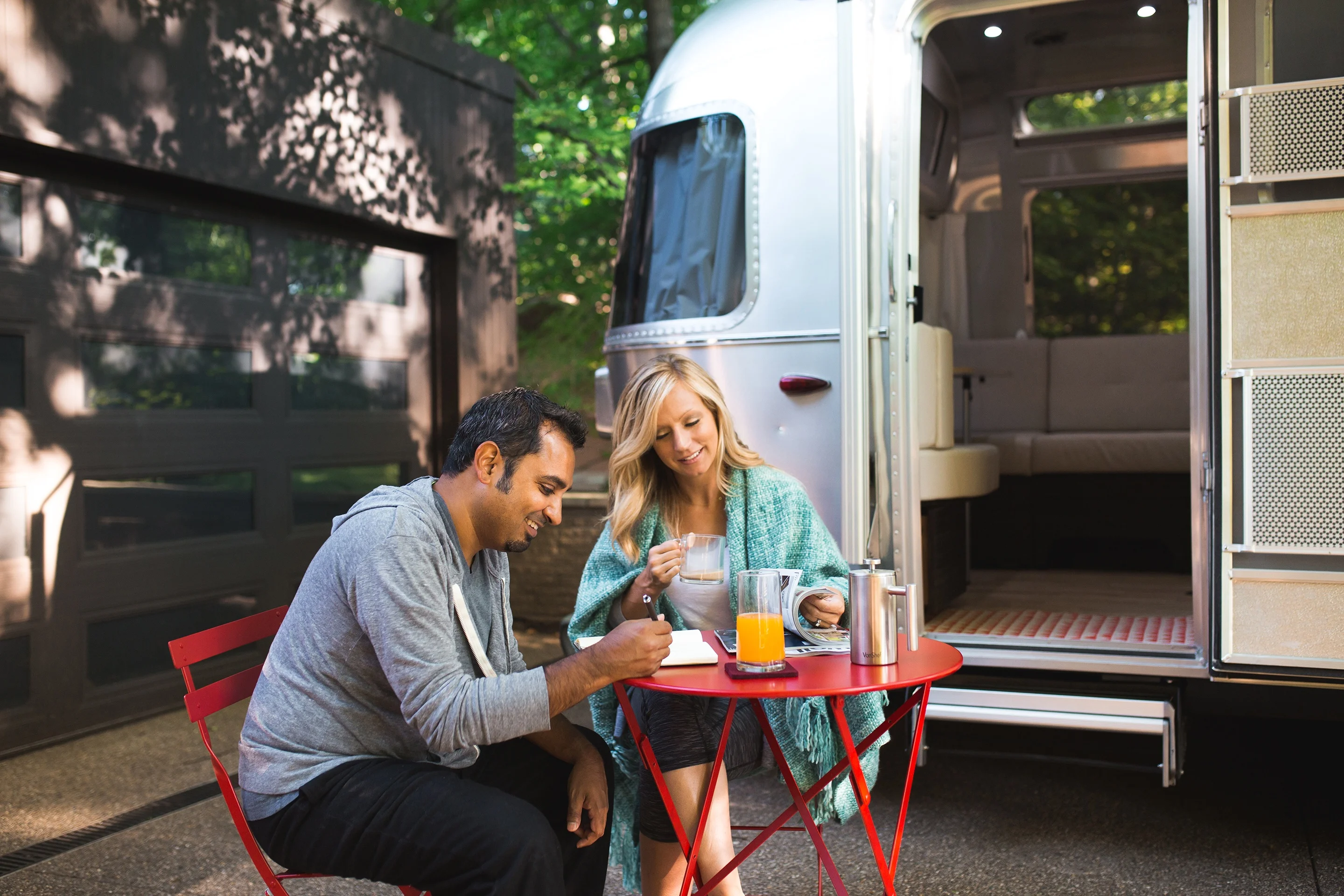 A couple enjoys coffee and juice at a red table outside their camper, with greenery in the background on a sunny day.