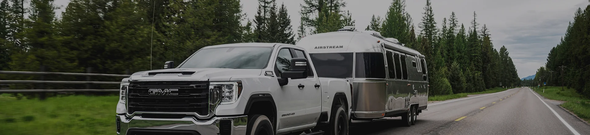 Pickup truck towing a large, silver camper on a forest-lined, rural road under a cloudy sky.