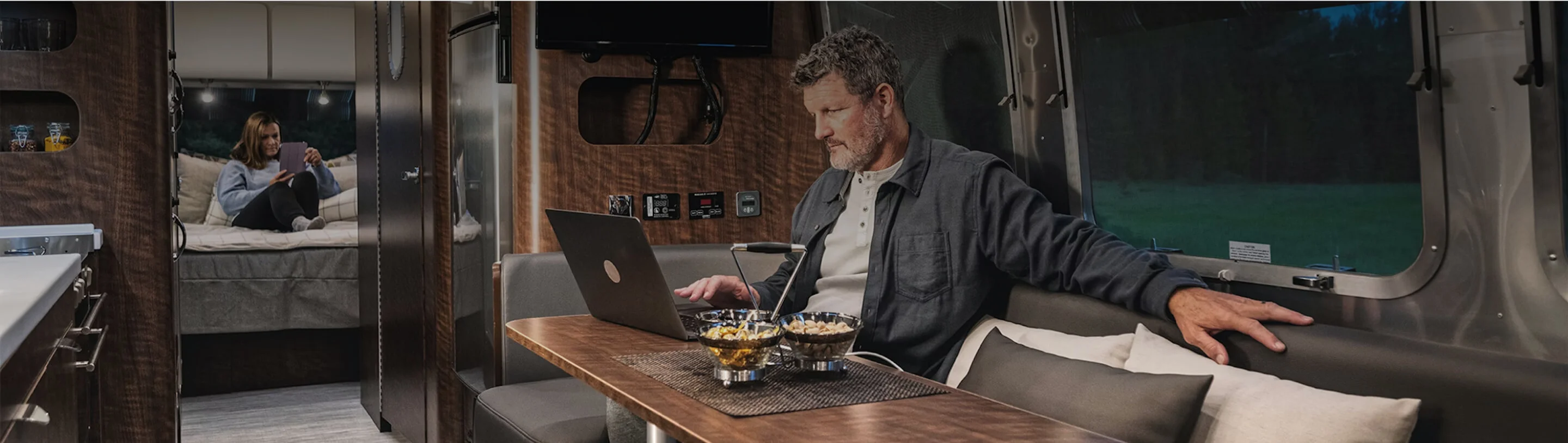 Man working on a laptop in an RV dining area, while a woman reads in the background. Cozy interior with cushions and subdued lighting.