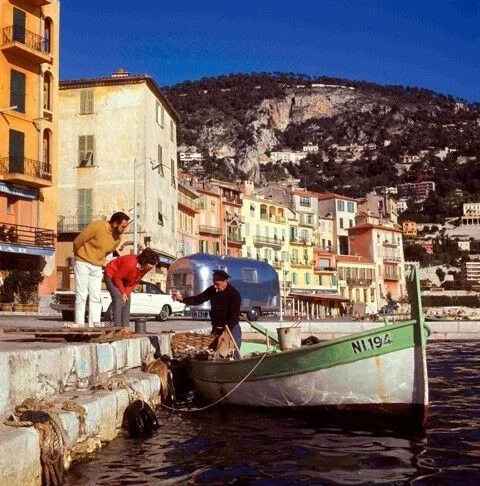 Two people on a colorful waterfront greet a fisherman on a small green boat. Behind them are vibrant buildings and a hillside with greenery, under a clear blue sky.