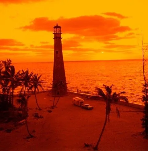 A lighthouse stands near the shore with palm trees and an RV nearby under a vivid orange sunset sky, overlooking a calm ocean.