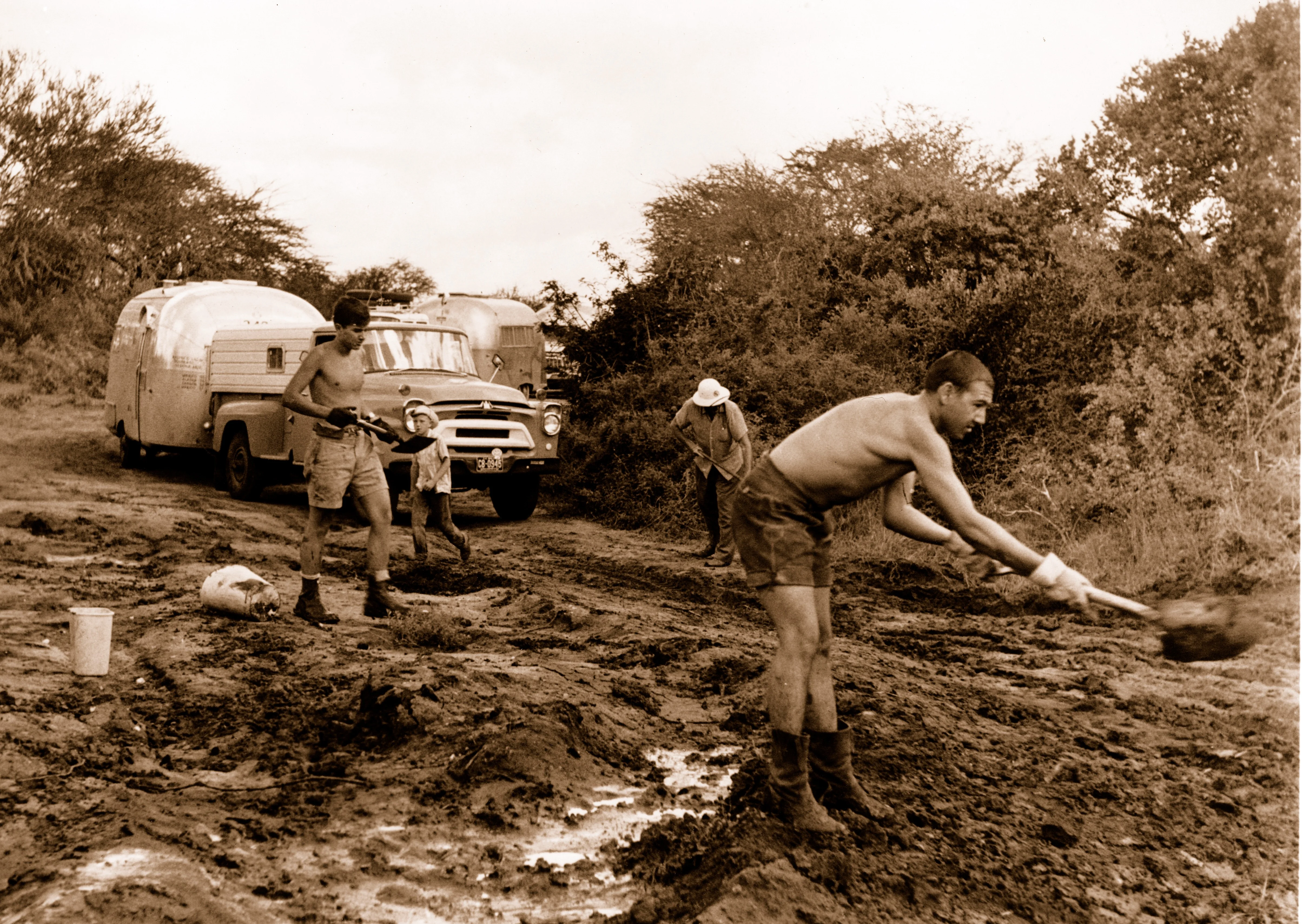 Four men work to clear mud on a rural road, shoveling earth beside a vintage truck and trailer, surrounded by trees.