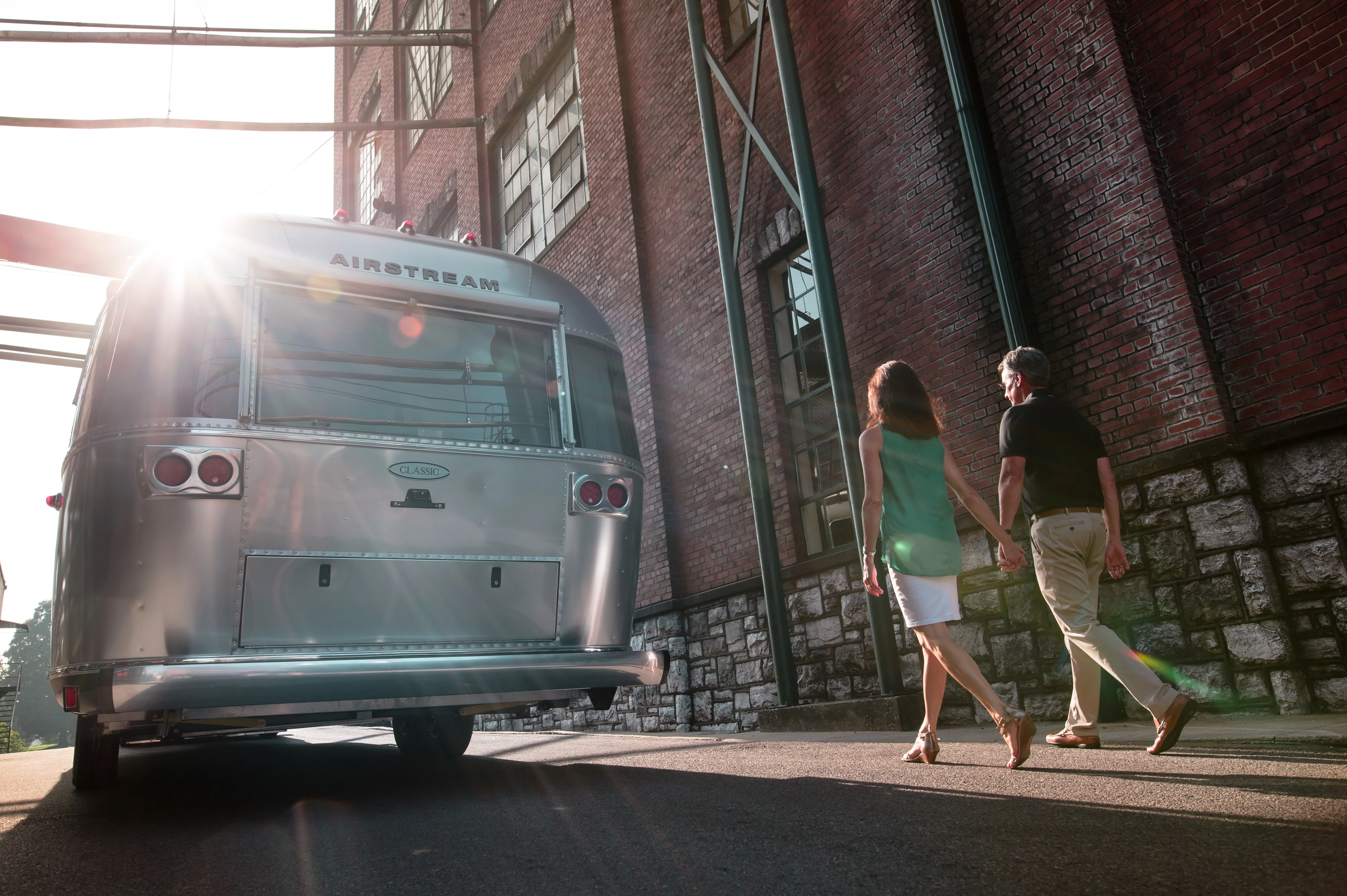 A couple holds hands while walking beside an Airstream trailer parked next to a brick building, with sunlight streaming through the scene.