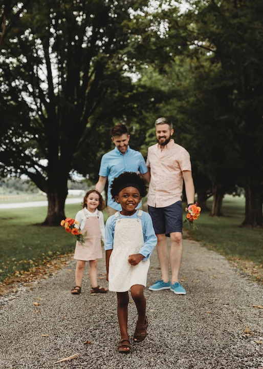 A joyful family walking down a tree-lined path, with two adults and two children smiling, holding bright flowers.