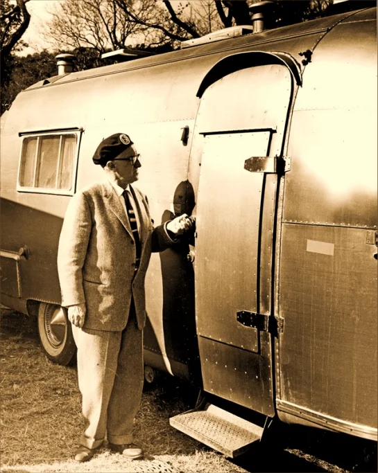 A man in a suit and beret stands beside an old trailer, holding its door handle, in a sepia-toned photograph.