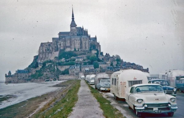 A row of vintage cars and trailers parked along a narrow road leading to Mont Saint-Michel, a historic island abbey in France, with overcast skies in the background.