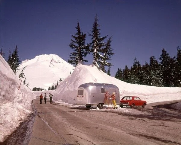 A snowy mountain road with tall snowbanks, a silver travel trailer, and a red car parked beside it. Three people stand near the trailer, and two more walk along the road. Pine trees and a snow-covered mountain are in the background.