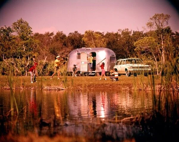 A family enjoys outdoor activities near a pond, beside a silver camper and a vintage car. Trees and grass surround the area, and the scene has a warm, nostalgic feel.