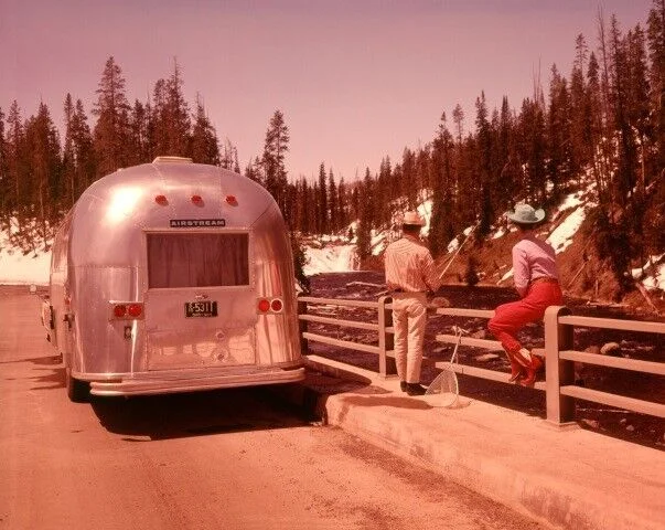 A shiny Airstream trailer is parked by a river. Two people stand on a snowy roadside, one leaning on the bridge railing, both looking at the water surrounded by pine trees.