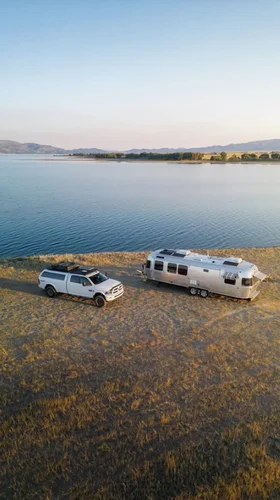 Aerial view of a white truck towing a silver camper beside a serene lake, with mountains in the background under a clear sky.